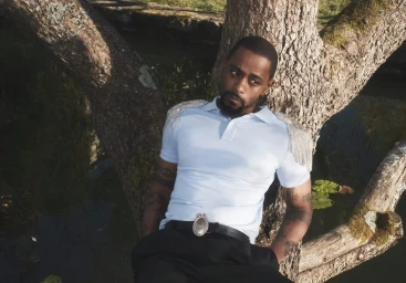 Lakeith Stanfield reclines against a mossy tree trunk wearing a light blue fringe-embellished polo shirt and black trousers.