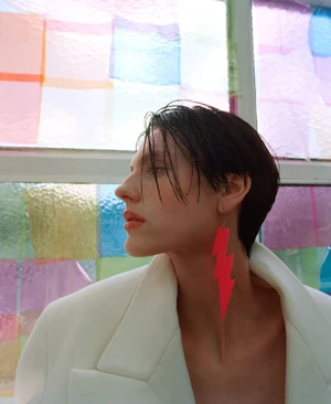 Profile portrait of a model with short dark hair wearing a tailored white blazer and a bright red lightning bolt earring.