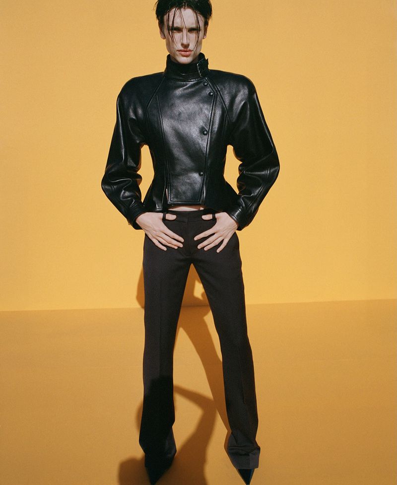Model Charlie Jones posing in a structured black leather jacket and black trousers against a warm yellow studio background.