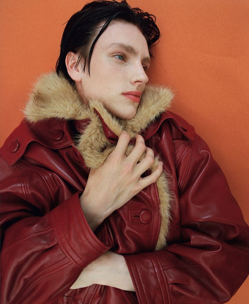 Charlie Jones wearing a burgundy leather shearling-trimmed coat against an orange backdrop in a fashion portrait.