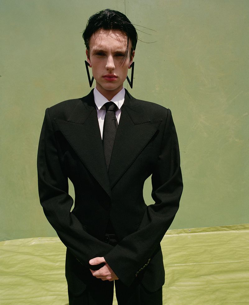 Model Charlie Jones wears an hourglass black tailored suit and oversized geometric earrings against a light green textured backdrop.