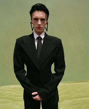 Model Charlie Jones wears an hourglass black tailored suit and oversized geometric earrings against a light green textured backdrop.