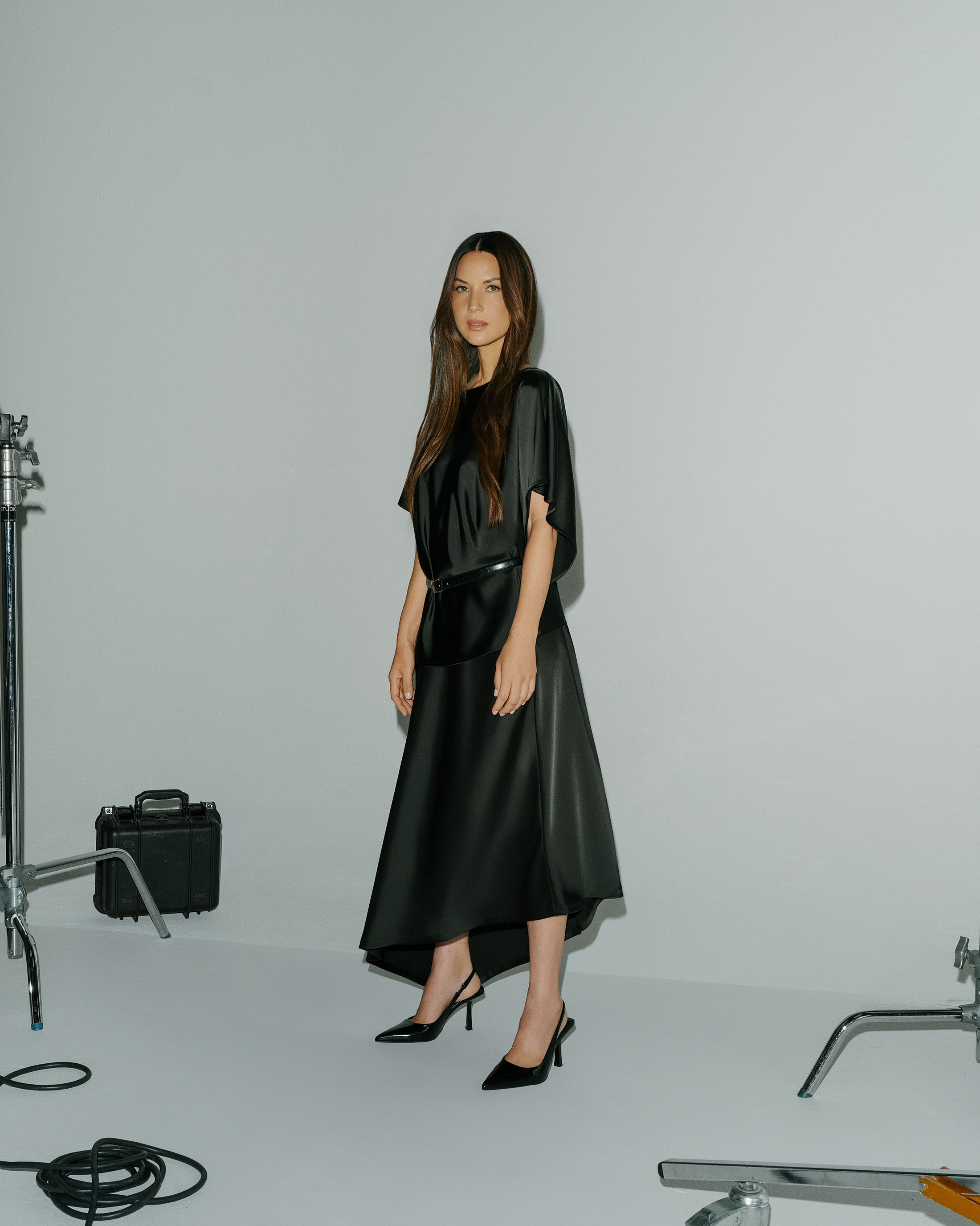 Olivia Munn in a black satin midi dress and black slingback pointed-toe heels posing in a studio with lighting equipment visible.