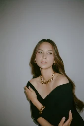 Olivia Munn wearing an off-the-shoulder black dress and chunky gold jewelry against a grey studio backdrop.
