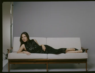 Olivia Munn reclining on a white mid-century modern sofa wearing a sleeveless black sequin dress.