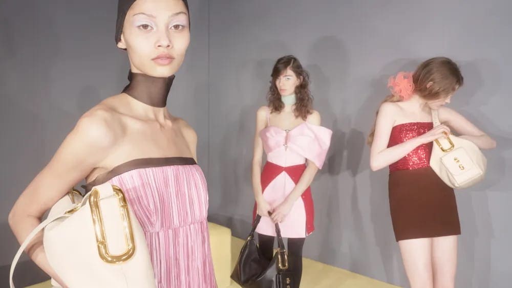 Three models posing against a grey wall holding Marc Jacobs leather shoulder bags with oversized gold hardware.