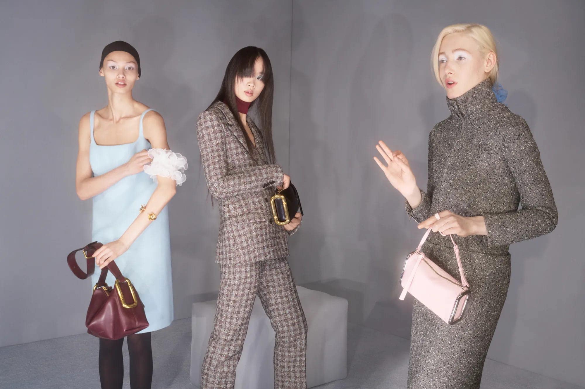 Three models presenting designer handbags while posing in tailored suits and a slip dress against a grey studio backdrop.