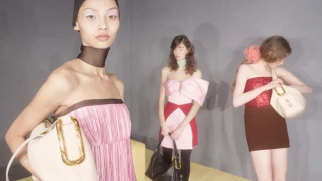 Three models showcase luxury handbags in pink, red, and brown designer dresses against a grey studio wall.