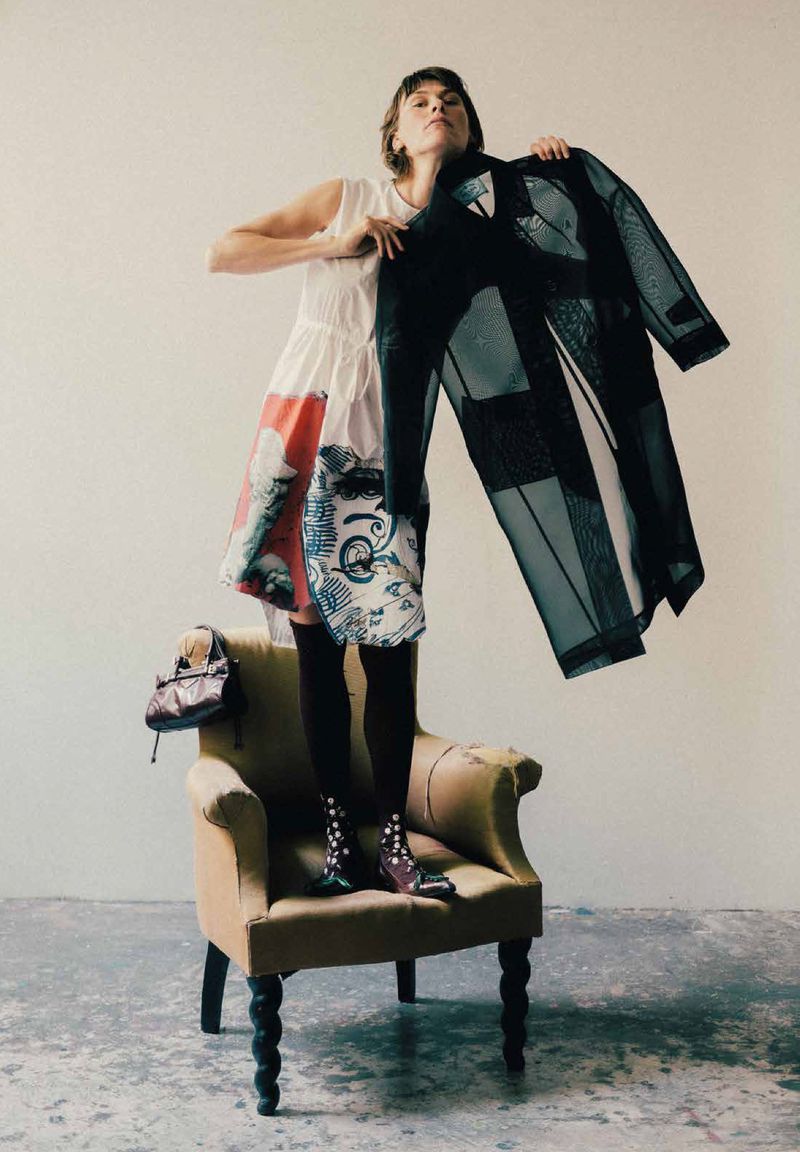 Fashion model standing on a vintage armchair holding up a sheer black coat against a neutral studio backdrop.