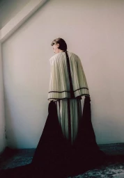 Model viewed from behind wearing a pleated cream capelet and skirt while holding a flowing black train in a minimalist room.