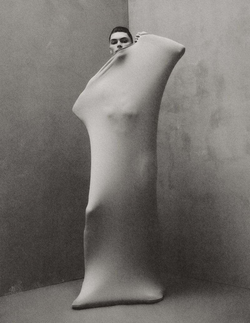 Monochrome portrait of a man partially concealed in a skin-tight garment in a corner studio, emphasizing form and texture.