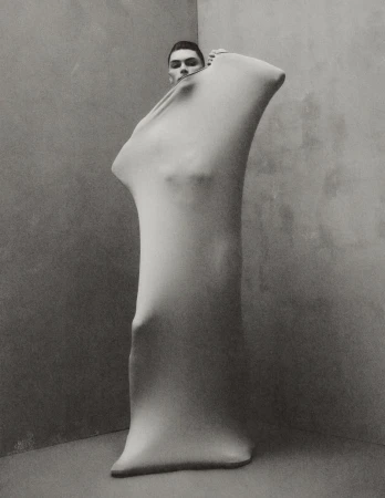 Monochrome portrait of a man partially concealed in a skin-tight garment in a corner studio, emphasizing form and texture.