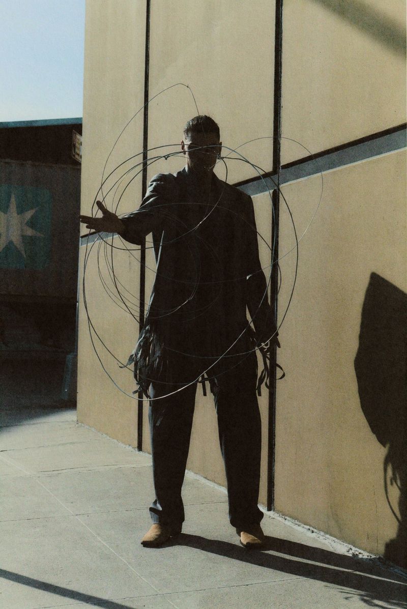 Adult male in a black jacket and pants stands outside a light tan building, encircled with thin metal rods, creating geometric forms.