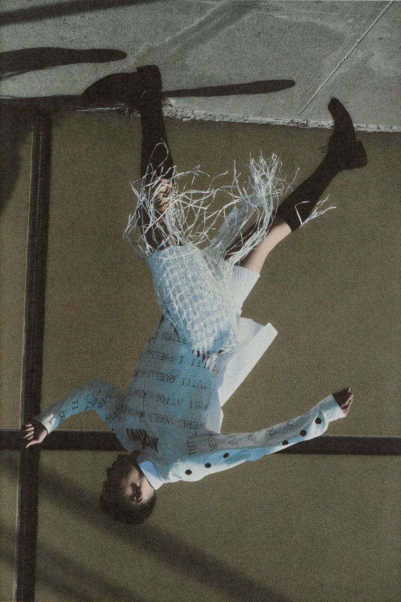 Fashion model suspended upside-down wearing a white printed long-sleeve top with fringed shorts and high black socks.