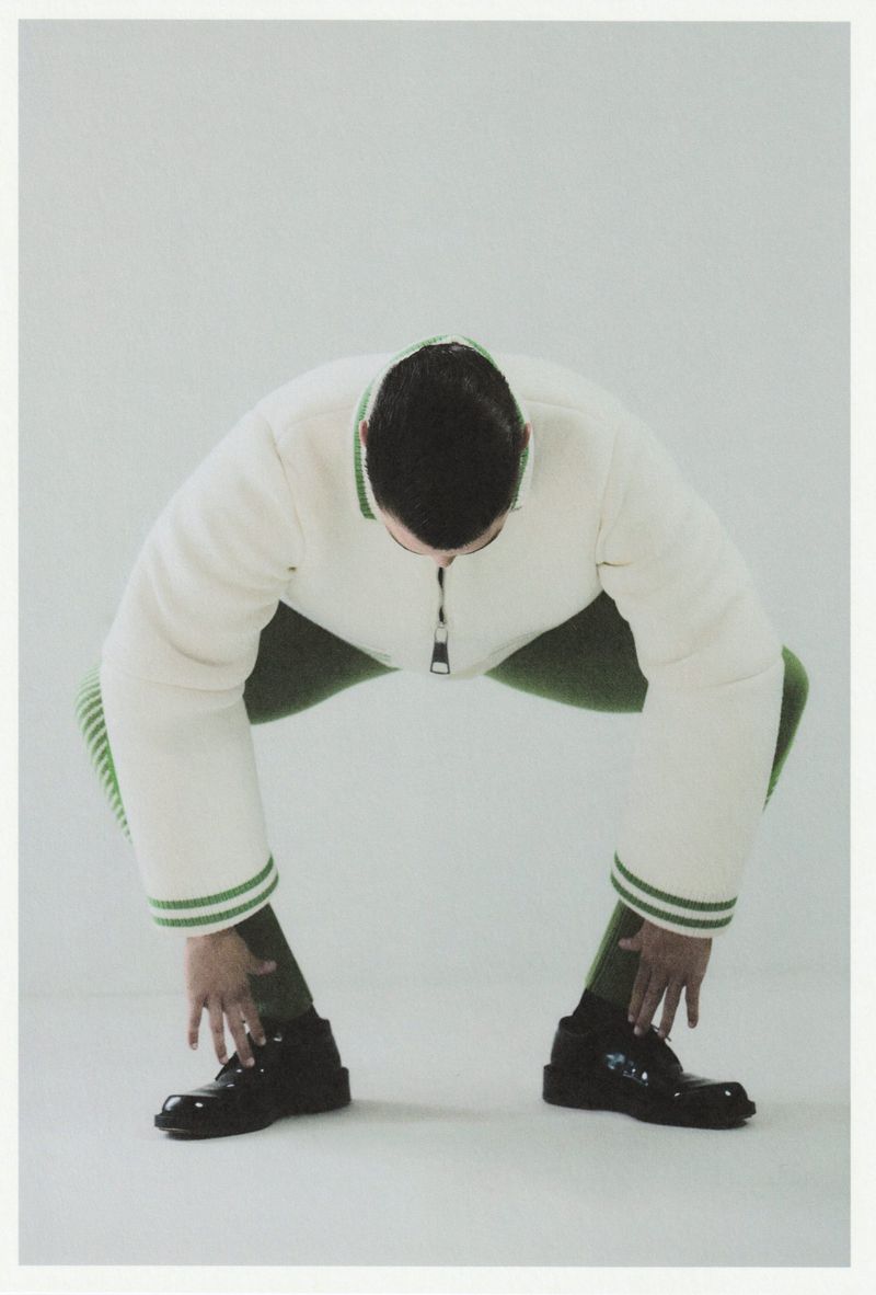 A person bending forward in an off-white jacket and green leggings touches their black shoes against a white background in a studio.