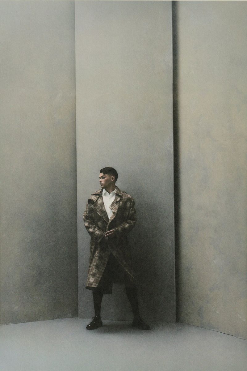Fashionable adult male model in an patterned trench coat, striking a pose indoors against a neutral grey backdrop, embodying modern style.