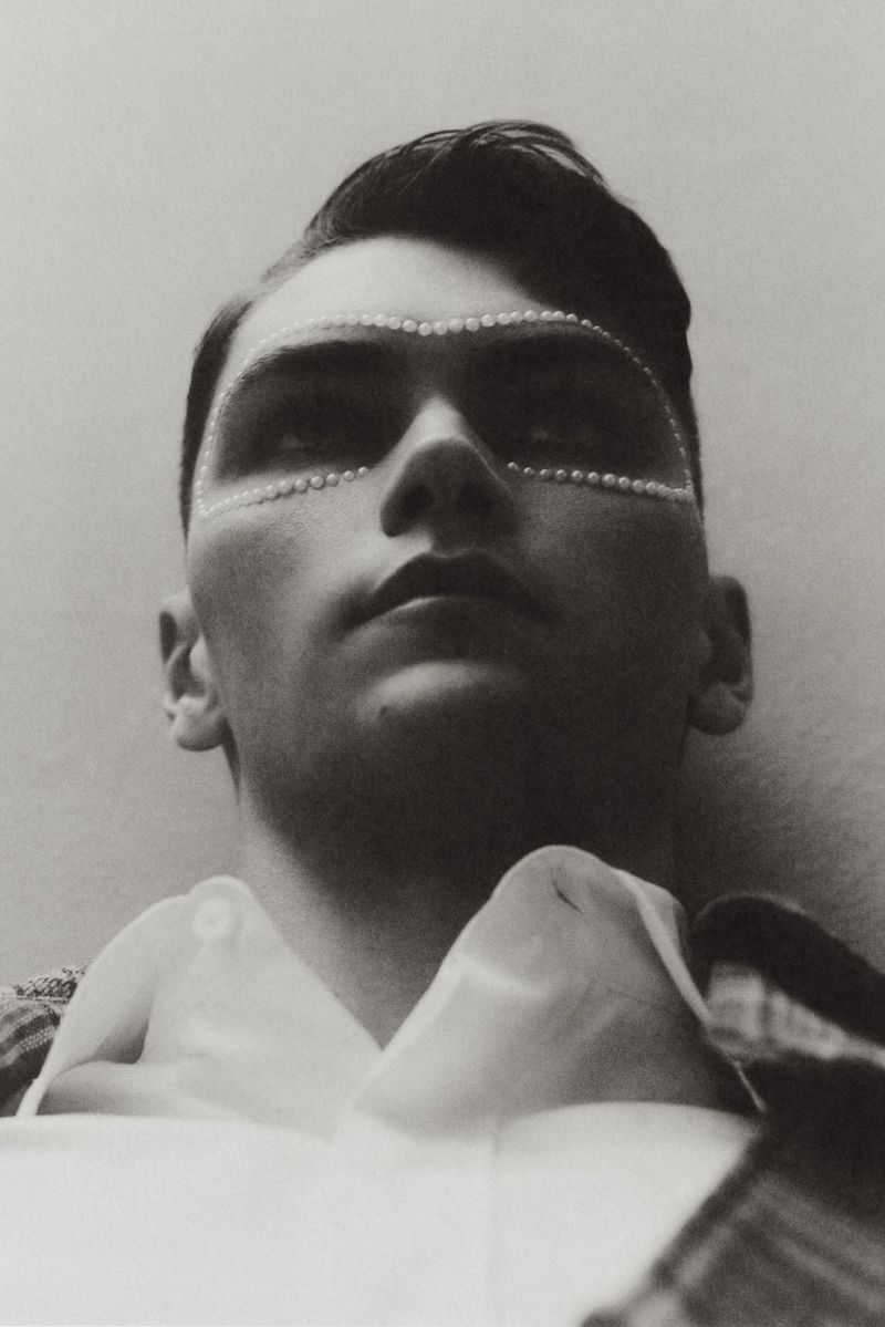 Close-up portrait of a man with pearl makeup around his eyes, looking upward with a neutral expression, in a studio with soft lighting.