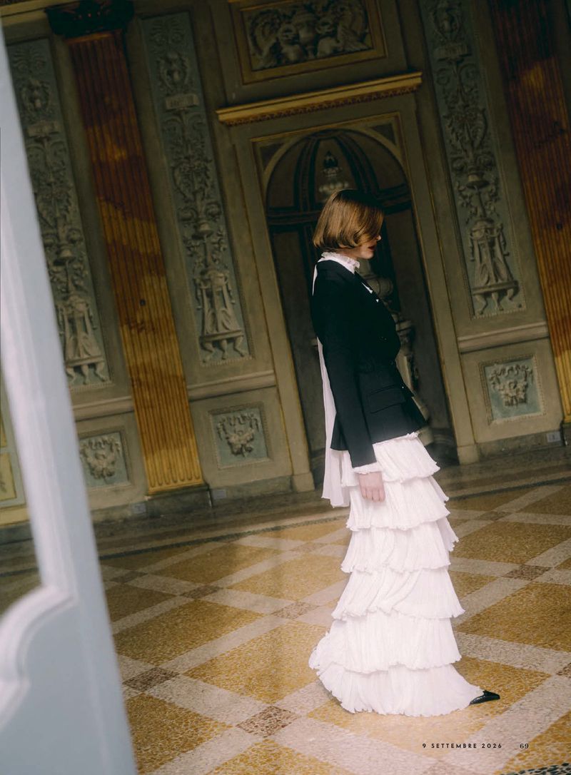 Model Hannah Weber in a black jacket and tiered white ruffled maxi skirt standing in an ornate classical hall.