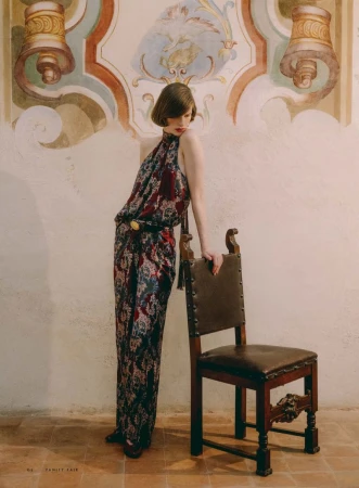 Model Hannah Weber wearing a patterned halter jumpsuit and leaning against an antique wooden chair before a fresco wall.
