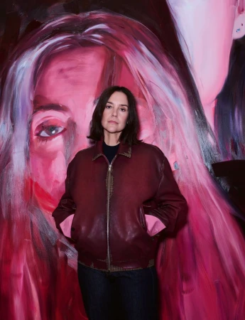 Woman in a burgundy leather jacket standing in front of an expressive pink painted portrait backdrop.