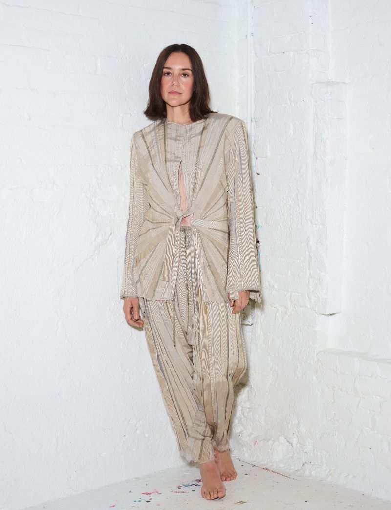 Woman standing barefoot in a textured beige matching jacket and trouser set against a painted white brick corner.