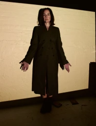 Woman in a long dark tailored coat standing with open palms against a rectangular beam of light on a textured wall.