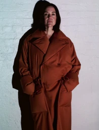 A woman wearing a voluminous rust-colored trench coat poses against a white painted brick wall casting a distinct dark shadow.