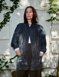 Woman in a paint-splattered dark denim chore coat standing against a white glass wall covered in climbing ivy.
