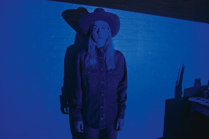 Model wearing a wide-brimmed western hat and denim shirt under deep monochromatic blue lighting with a sharp shadow.