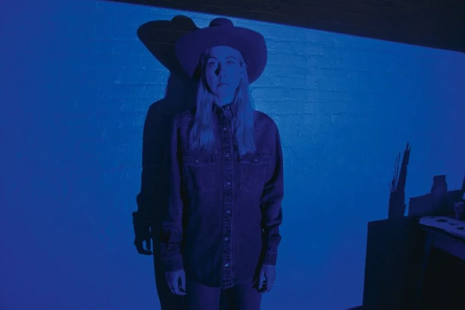 Model wearing a wide-brimmed western hat and denim shirt under deep monochromatic blue lighting with a sharp shadow.