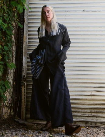 Blonde woman wearing an oversized dark draped outfit and wide-leg trousers standing in front of a closed metal roller shutter.