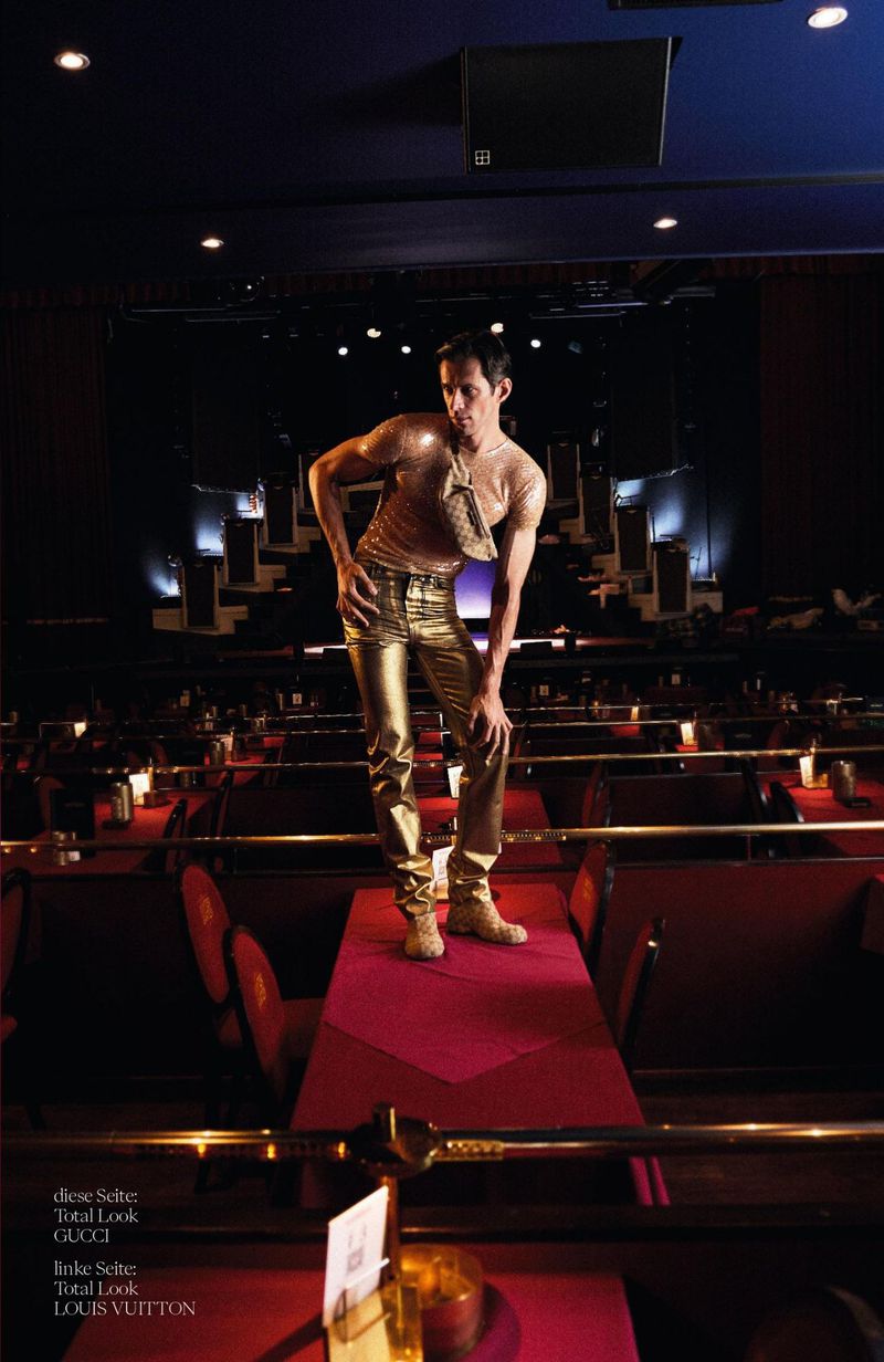 Model Kamil Skłodowski standing on a table in metallic gold trousers and a sheer top in a dimly lit auditorium.
