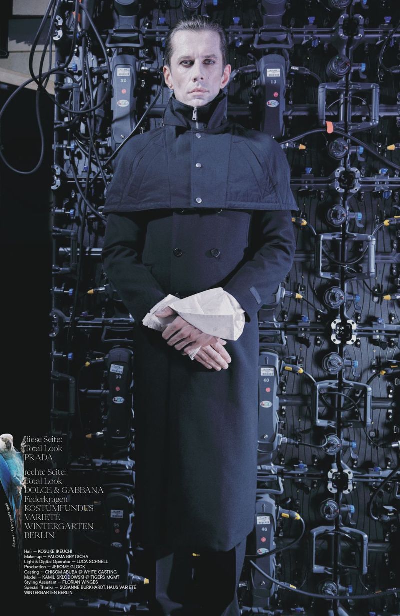 Model Kamil Skłodowski wearing a dark structured cape coat with pale cuffs against a stage lighting rig.