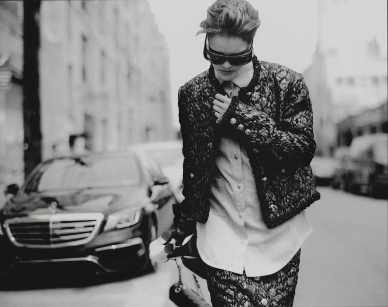 Model Amelie Sante walks along a city street wearing dark sunglasses, a textured jacket, and a button-down shirt near a parked car.