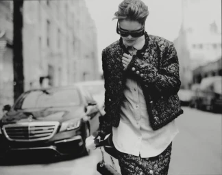 Model Amelie Sante walks along a city street wearing dark sunglasses, a textured jacket, and a button-down shirt near a parked car.