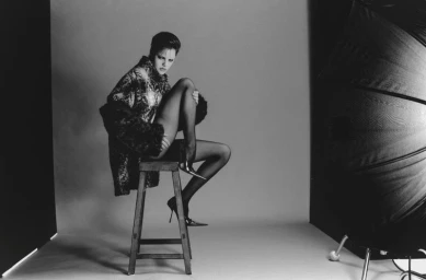 Black and white fashion portrait of model Amelie Sante seated on a wooden stool wearing a patterned coat and pointed high heels.