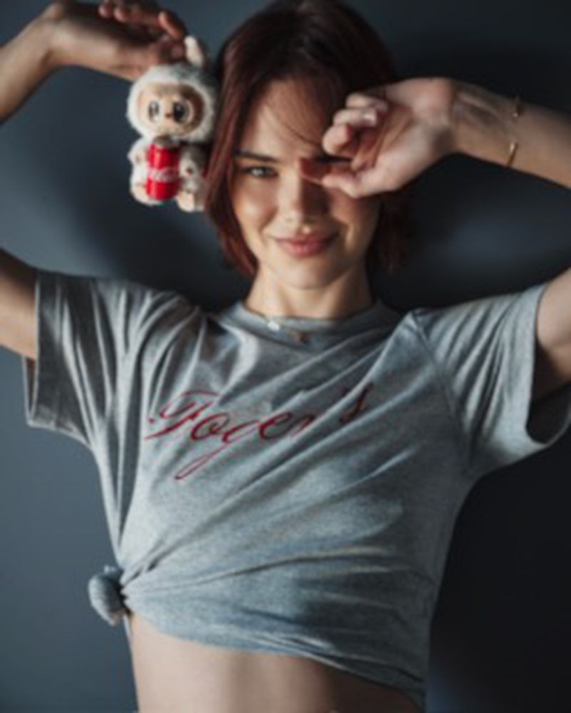 Woman in a tied grey t-shirt holding a small plush toy beside her head against a dark studio background.