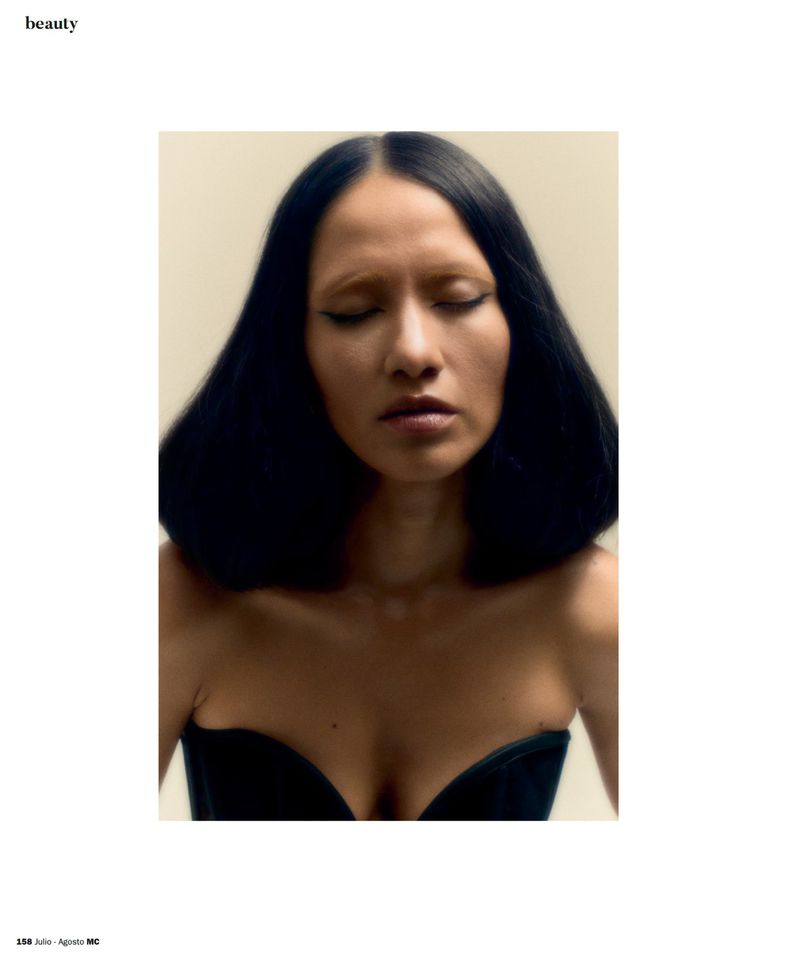 Portrait of a woman with voluminous dark bob hair, closed eyes, and light eyebrows in a black corset against a neutral backdrop.