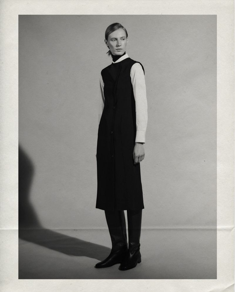 Model Julia Nobis standing in a black sleeveless midi dress and leather boots in front of a studio backdrop.