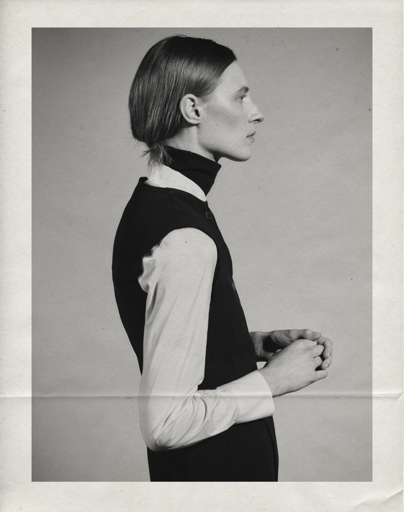 Model Julia Nobis in profile wearing a layered black vest and white long-sleeve shirt against a studio backdrop.