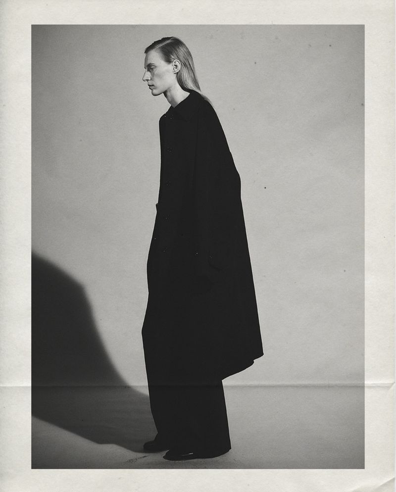 Full-length profile of model Julia Nobis wearing an oversized black coat and matching trousers in a monochrome studio setting.