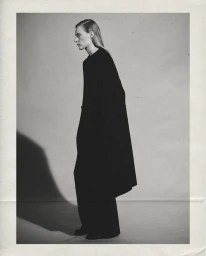Full-length profile of model Julia Nobis wearing an oversized black coat and matching trousers in a monochrome studio setting.