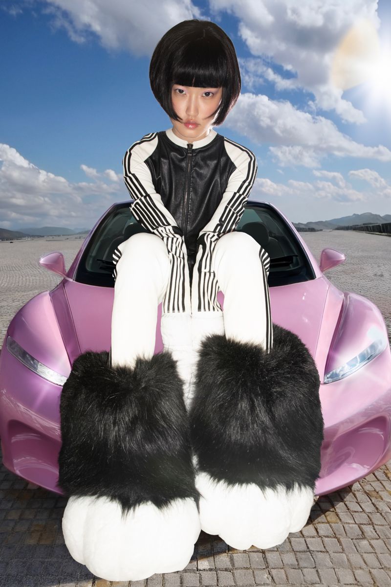 Young Asian woman with bob haircut sits atop a pink sports car in oversized, furry black-and-white monster-paw slippers against a blue sky.