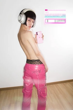 Non-binary fashion model poses in pink PVC pants and lace, wearing headphones and holding pink phone in a studio setting.