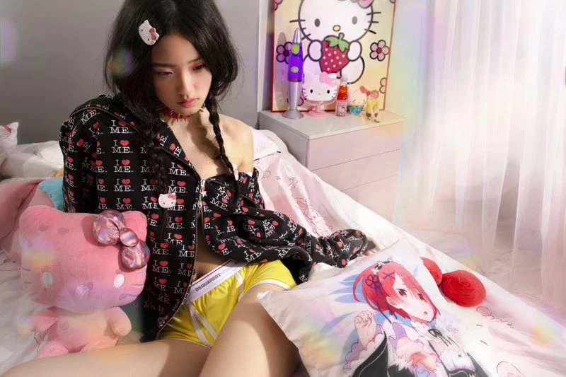Model Yirou Zhou sitting on a bed wearing an unzipped black graphic hoodie and yellow shorts surrounded by plush toys.