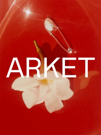 White flower and a silver safety pin resting in red liquid with bold white ARKET typography overlay.