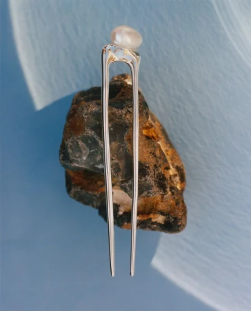 Silver two-pronged hair pin topped with a single pearl resting vertically against a textured rock on a light blue background.