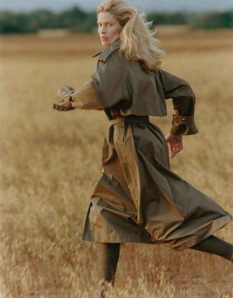 Woman with blonde hair wearing an olive green belted trench coat running through a golden wheat field and looking back.
