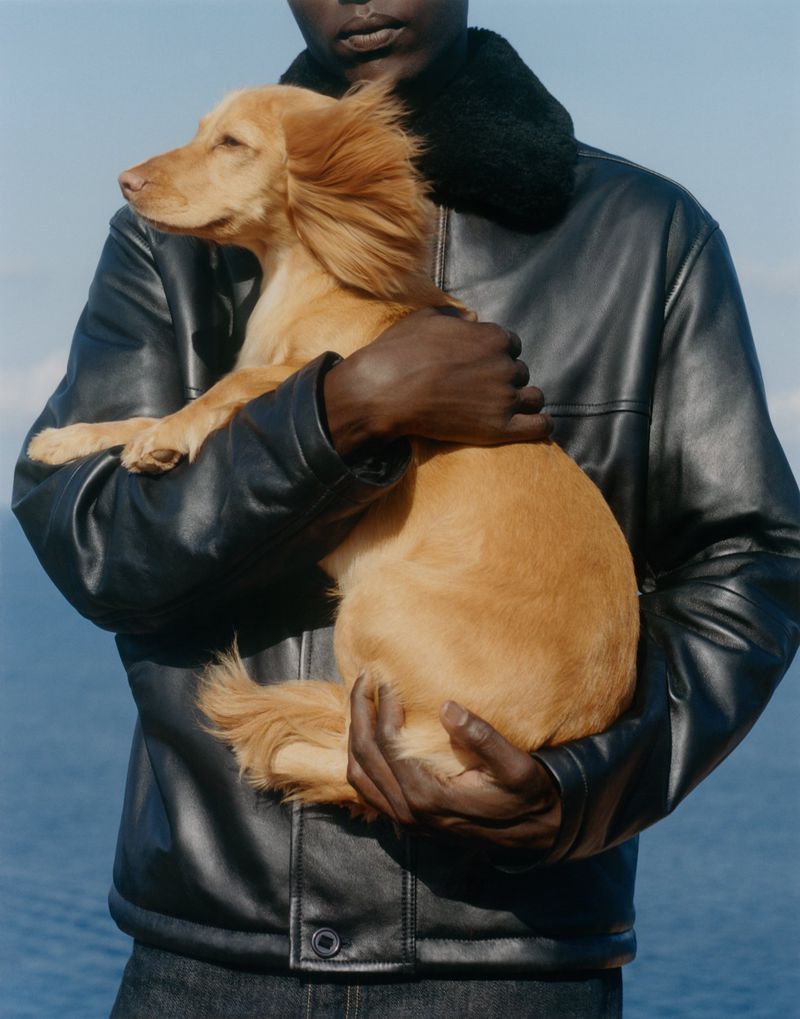 Black model wearing a black shearling-collar leather jacket holding a long-haired golden dog against a blue ocean background.