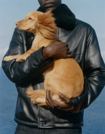 Black model wearing a black shearling-collar leather jacket holding a long-haired golden dog against a blue ocean background.